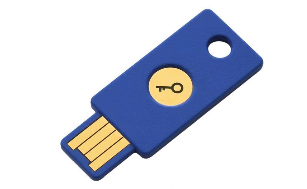 A blue USB security key with a gold circular emblem featuring a key symbol. The USB connector is exposed at one end, and the device has a hole near the opposite end, possibly for attaching to a keychain.