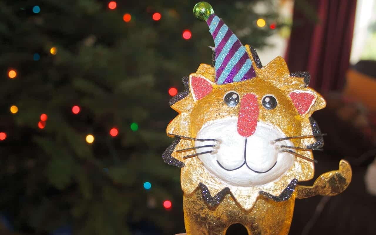A festive, glittery lion ornament with a cheerful face and wearing a blue and purple striped party hat is in the foreground. In the background, there are colorful Christmas lights and a blurred Christmas tree.