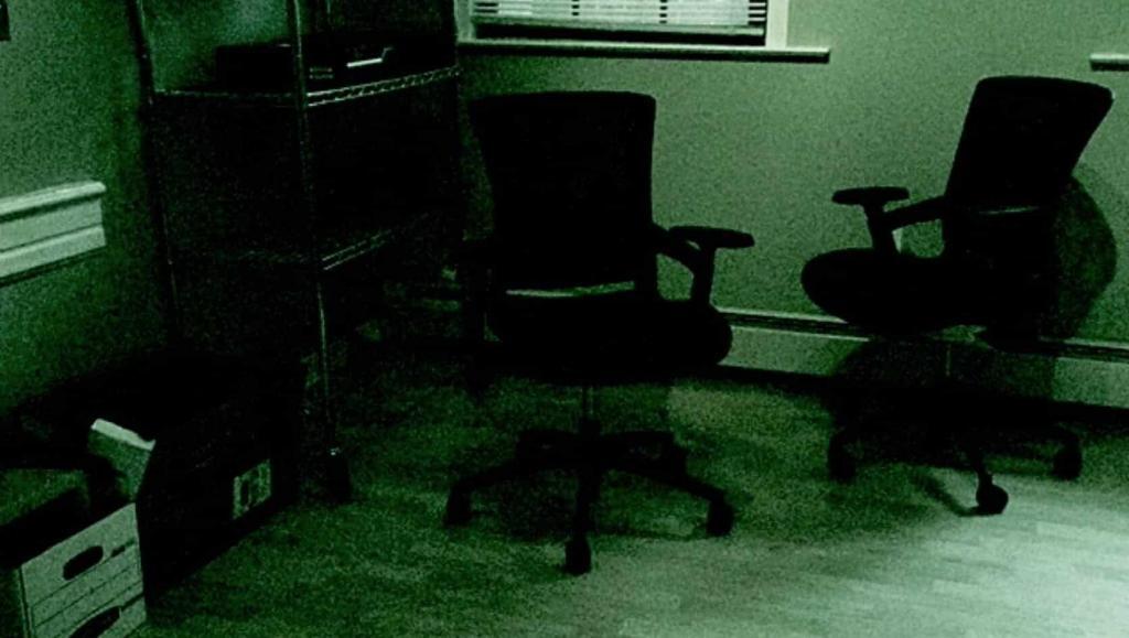 A dimly lit room with two empty office chairs facing each other. There is a metal shelving unit on the left side with boxes and papers, and a window with closed blinds on the wall. The overall scene has a greenish tint.