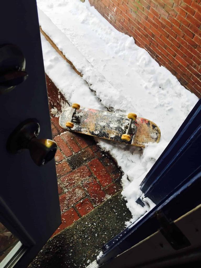 A skateboard lies on a snow-covered brick pathway just outside an open blue door. Snow and ice are scattered across the ground.