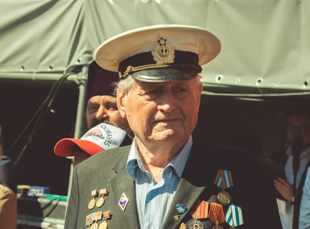 An elderly man in a naval uniform adorned with various medals and badges stands outdoors. He wears a white cap with an emblem and looks into the distance. He seems to be at a public event, with other people partially visible in the background.