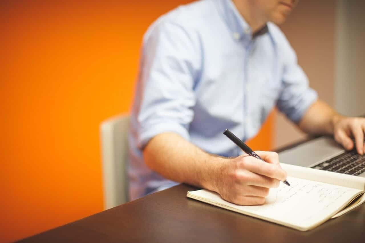 A tech MSP managed consultant in a light blue shirt writes in a notebook with a pen while using a laptop, seated at a desk with an orange wall in the background.