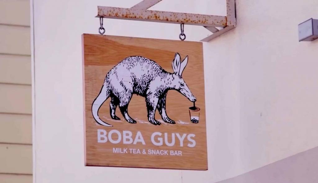 A wooden sign for "BOBA GUYS Milk Tea & Snack Bar" hangs outside, featuring a black-and-white illustration of an aardvark drinking from a cup with a straw—sure to catch the eye of any tech MSP managed consultant passing by.
