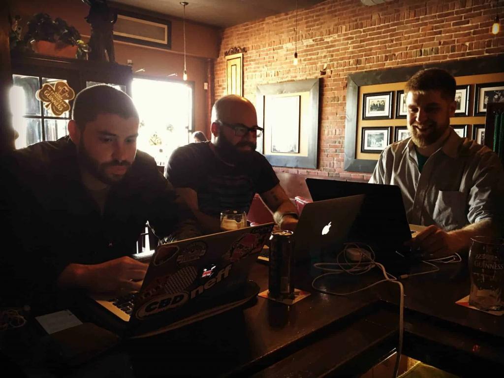 Three men sit at a dark wooden table in a cozy, brick-walled cafe, working as tech consultants on laptops. Sunlight streams in through the window, and drinks are placed beside them, creating a collaborative managed MSP atmosphere.