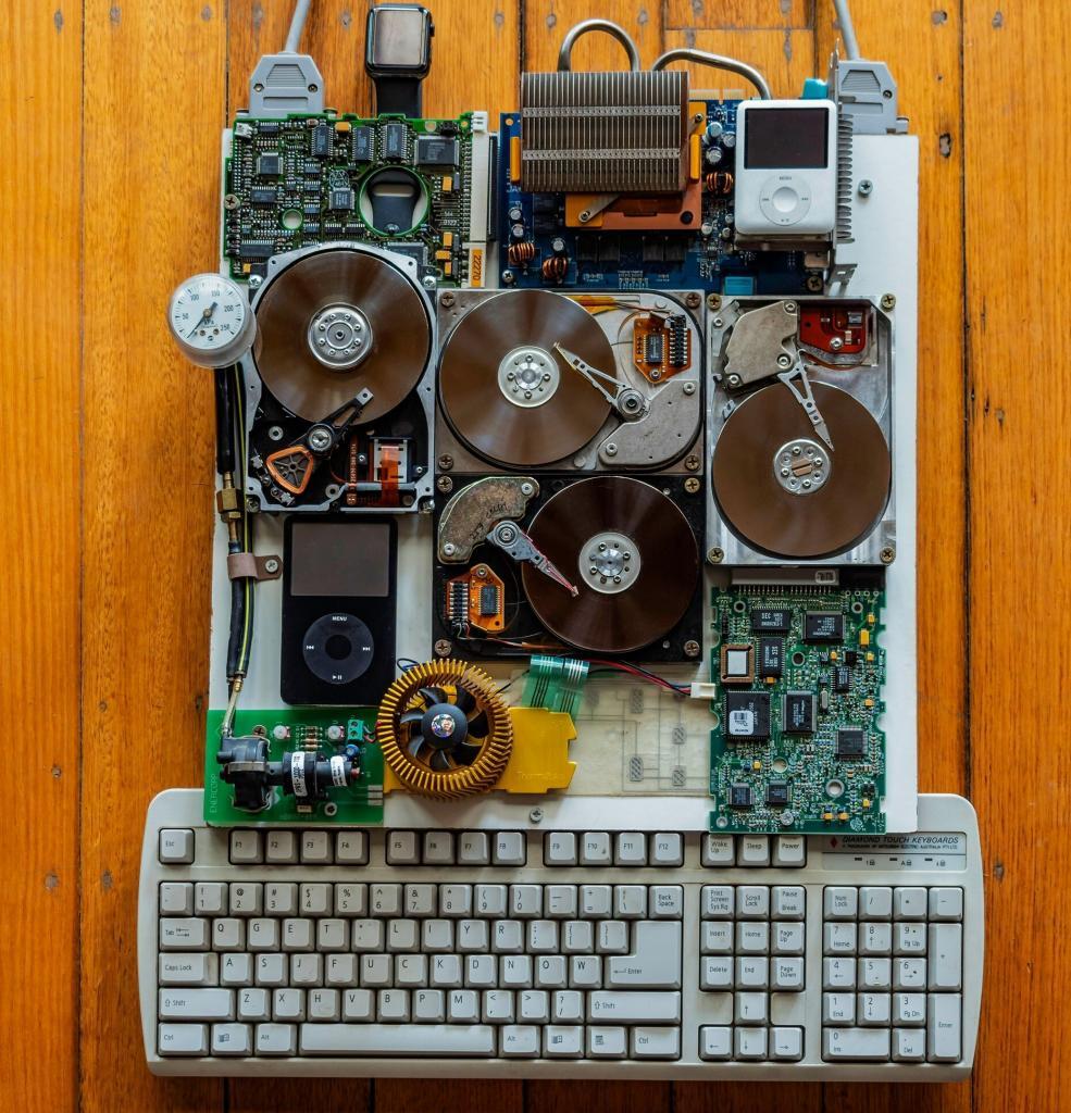 A vintage computer keyboard is surrounded by various electronic components, including circuit boards, spinning hard drives, wires, and an old iPod—reminiscent of a tech MSP managed consultant’s toolkit—arranged on a wooden floor.